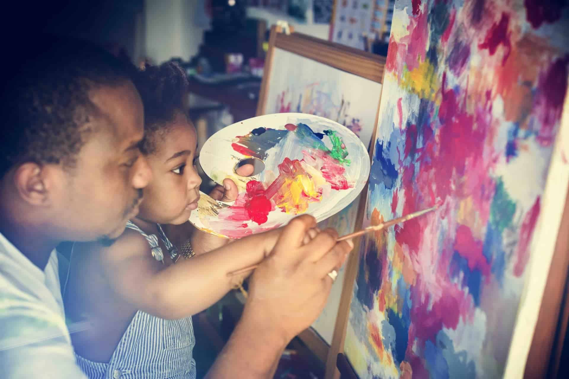 African descent dad teaching his child how to paint A father helps his toddler with her painting, representing the care that integral ecology requires to be focused upon the human person