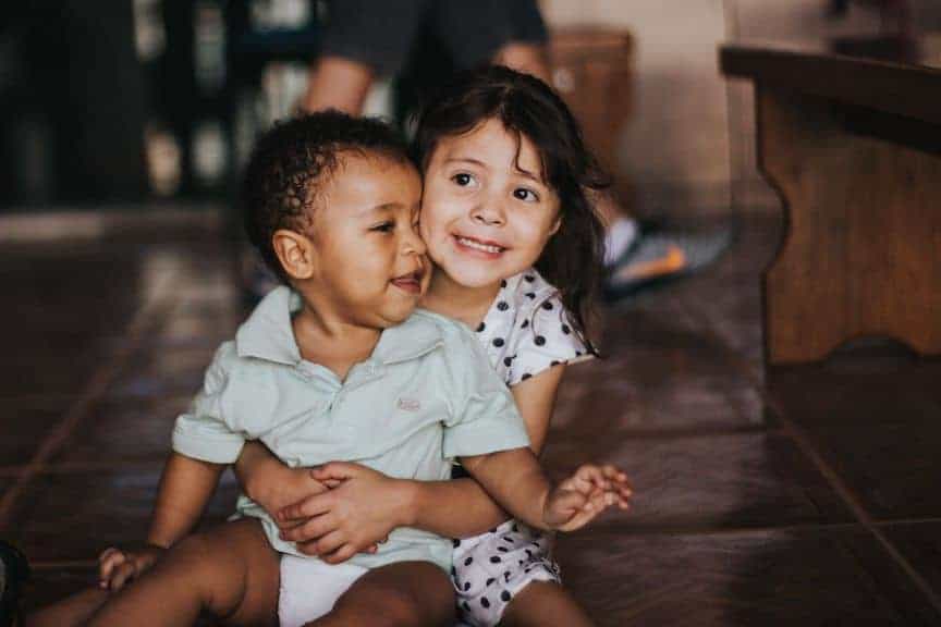 children-siblings-brother-sister-hug-laugh-smile-family-dignity-child A young girl sitting on the floor hugs a younger boy, calling to mind human dignity, the first of the three principles of catholic social teaching
